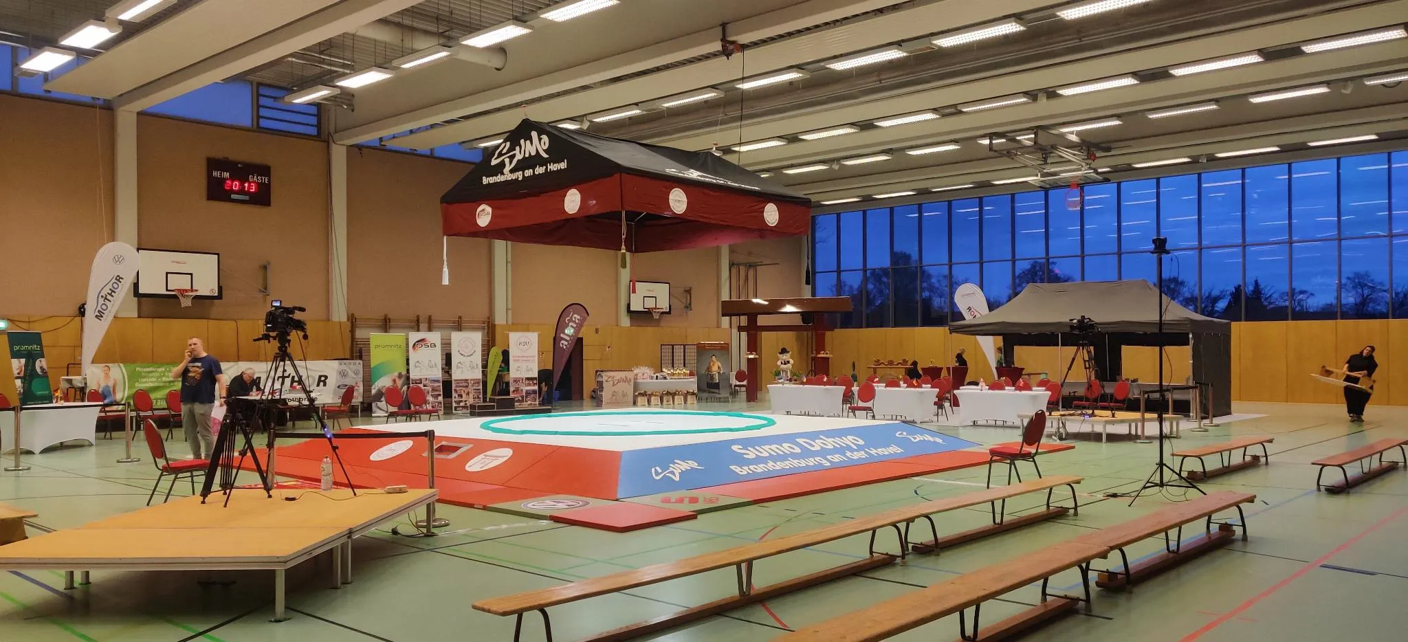 Photo du Dohyo, légèrement surelevé sous un tsuriyane en tissu au milieu d'un gymnase vide, et sur les côtés 2 caméras sur pieds pointés vers le centre