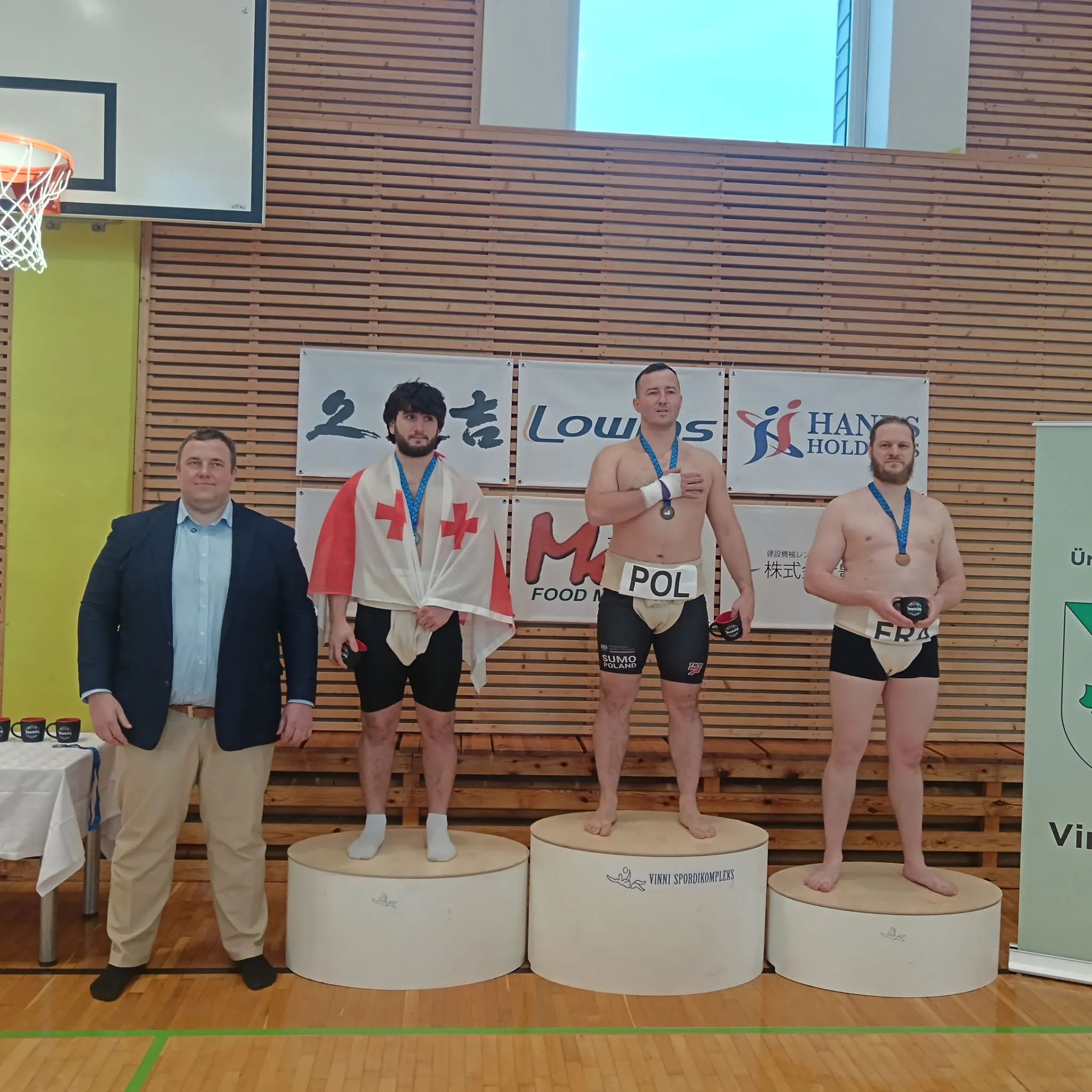 Photo d'un Podium avec 3 hommes en mawashi. Sur le mawashi à la première : POL. En 2è
position: GEO. En 3è: FRA. Sur le coté d'une podium, un homme de grande carrure en costume.