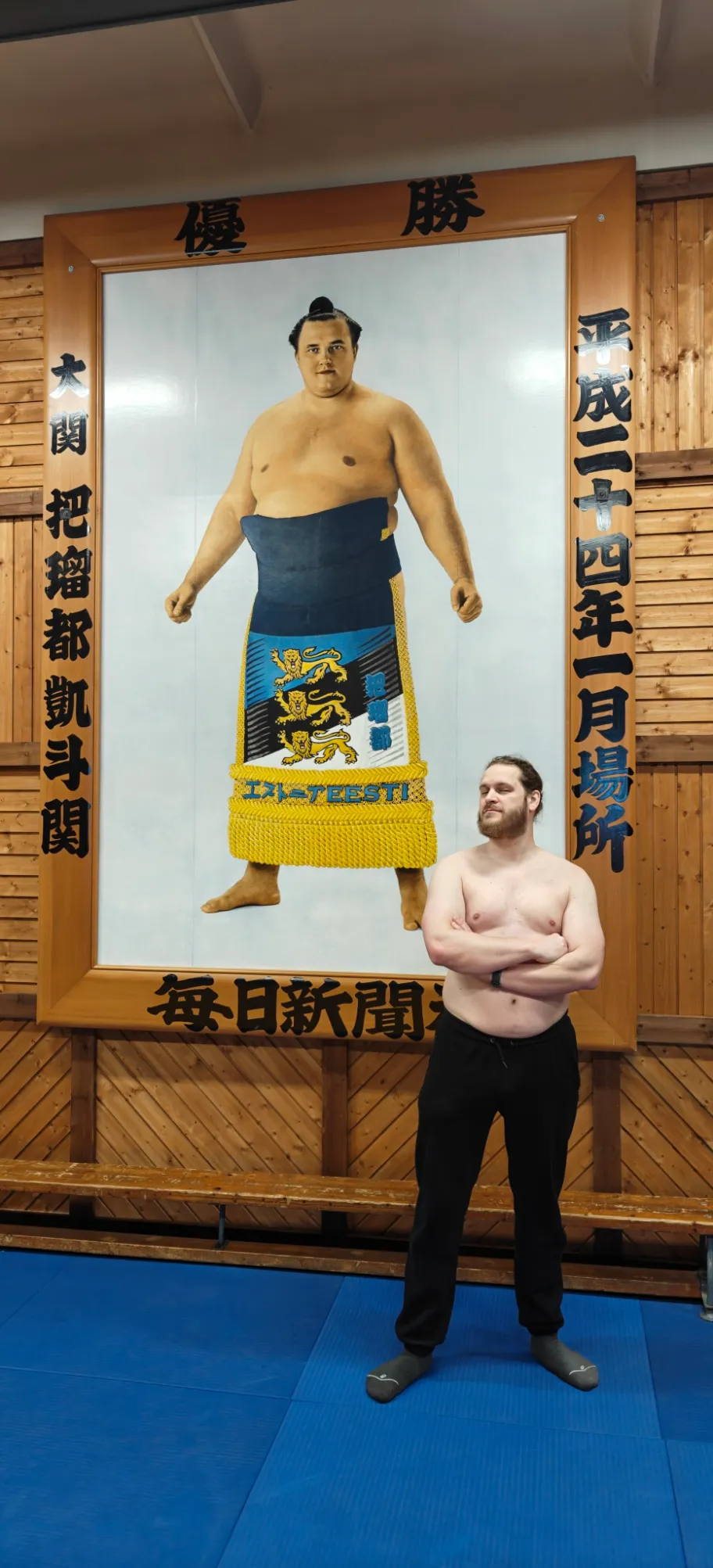 Photo d'un homme torse nu et bas de survêtement posant devant un portrait d'environ 2m
de haut sur lequel on peut avoir un homme ceint d'un keshō-mawashi composé d'un drapeau estonien sur lequel sont
présents 3 lions léopardés et sur lequel on peut lire 'EESTI'. Le portrait est entouré d'un cadre avec des
inscriptions en japonais.