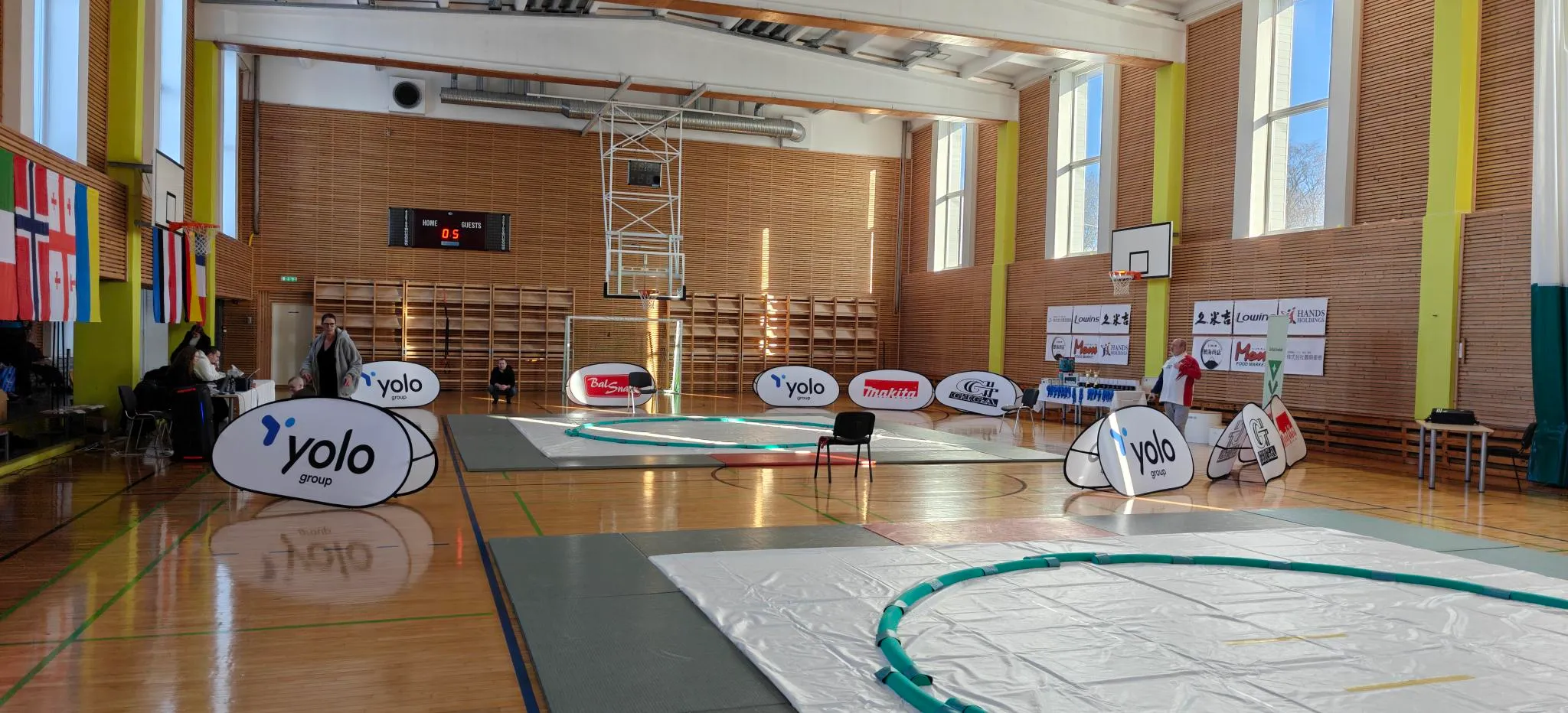 Photo d'un Gymnase où l'on voit 2 dohyo, des drapeaux sur le mur de gauche, des sponsors sur
le mur de droite, ainsi qu'au sol. Quelques personnes sont visibles sur la photo vacant à leurs occupations